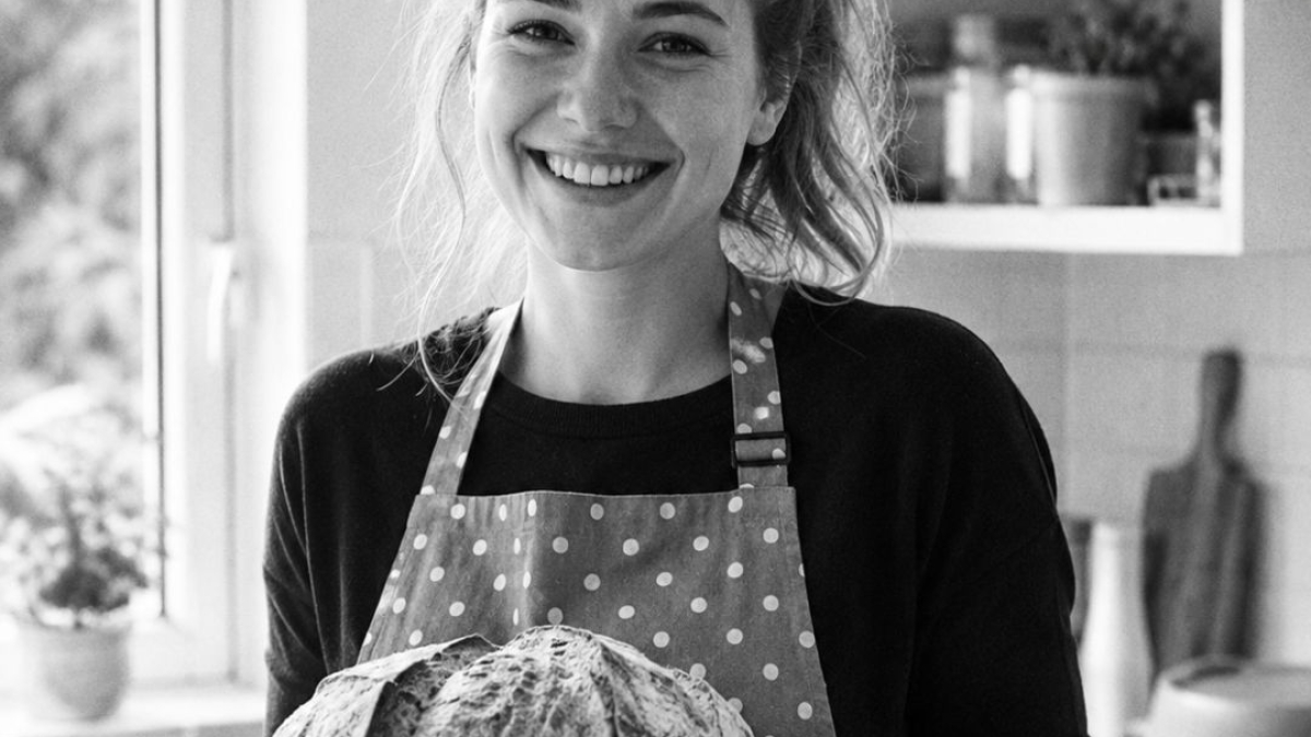 Jeune femme en tablier à pois tenant un pain fait maison dans une cuisine rangée, image en noir et blanc